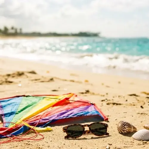 A close-up, sun-drenched photograph on a Puerto Rican beach featuring a colorful child’s kite resting on the sand next to a pair of small toddler sunglasses and natural seashells. The ocean is in the background.